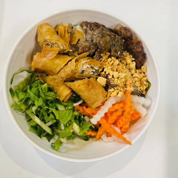 A white bowl filled with Vietnamese vermicelli noodles, grilled beef, crispy spring rolls, and fresh vegetables.