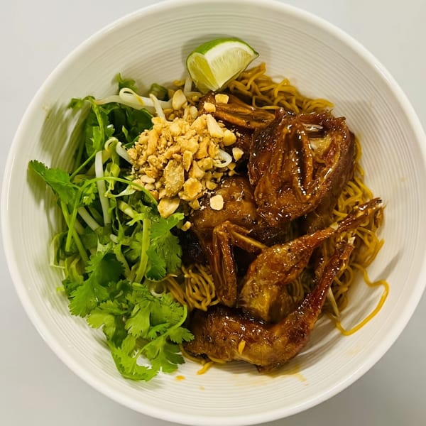 A bowl of noodles with glazed quail, topped with crushed peanuts, cilantro, bean sprouts, and a lime wedge.
