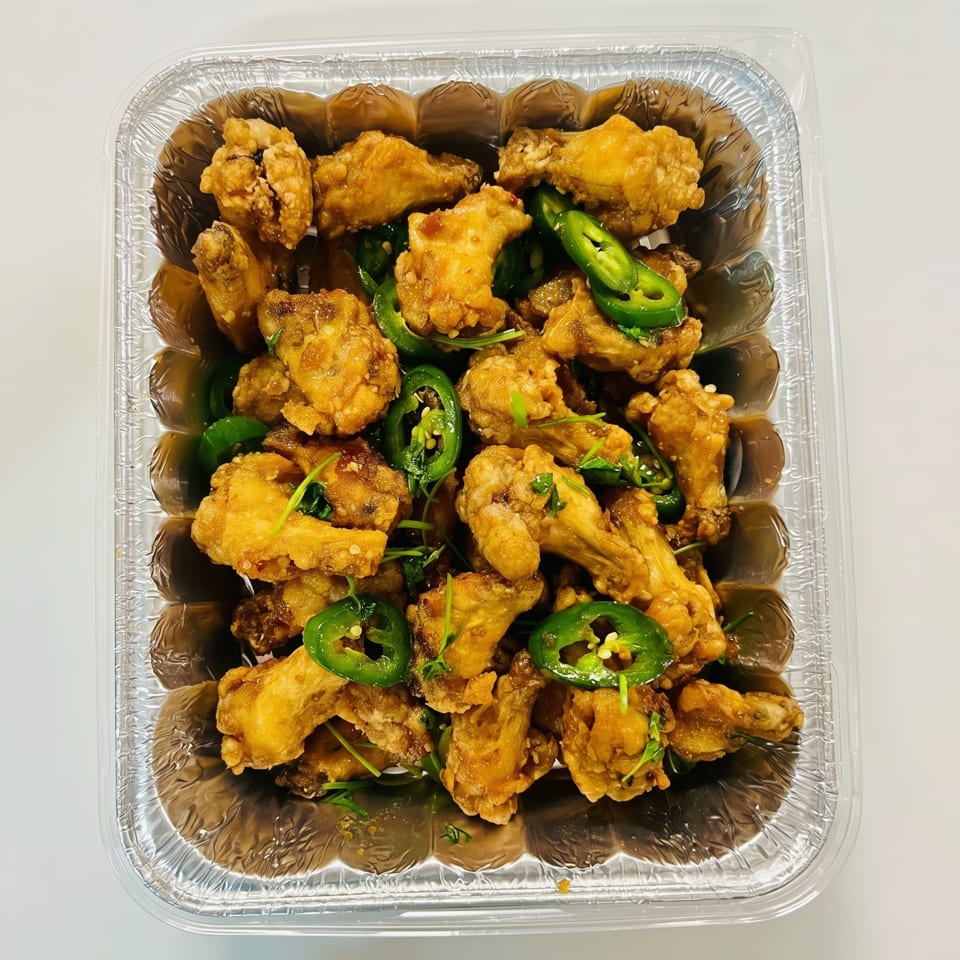 Crispy fried chicken wings with sliced green jalapeños and herbs in a foil takeout container.